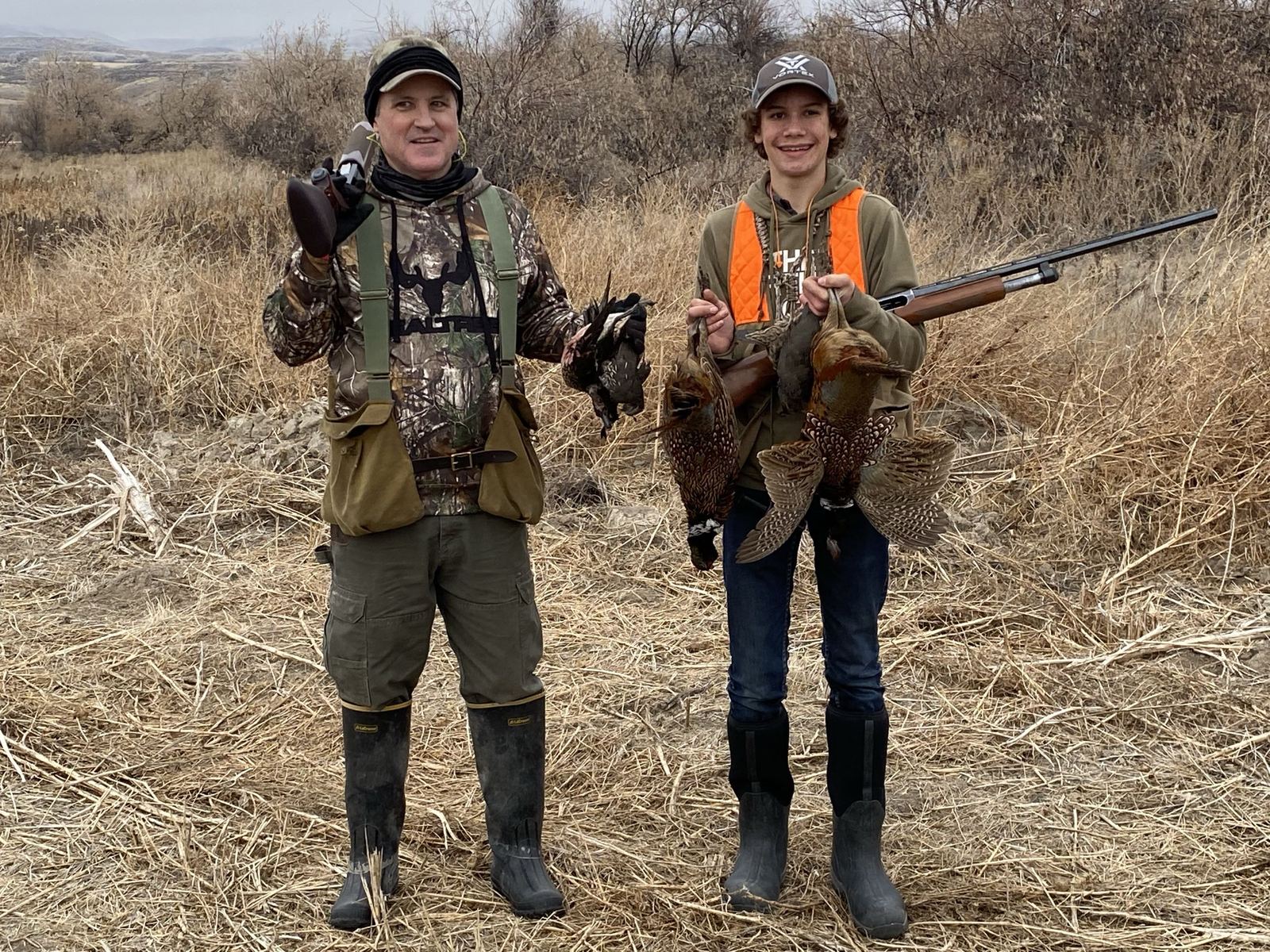 A father and son in hunting gear holding harvested birds after a day hunting the ranch