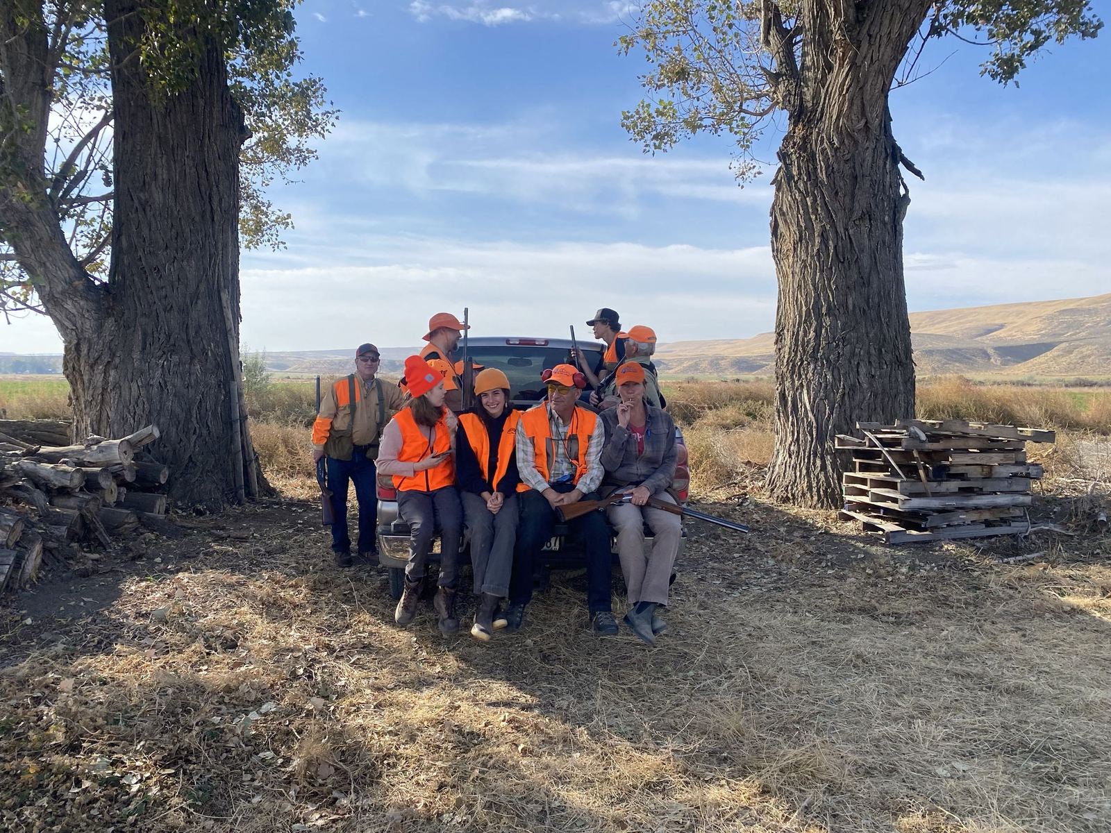 A small hunting party in orange vests gathered on a truck tailgate between two large cottonwood trees on the ranch